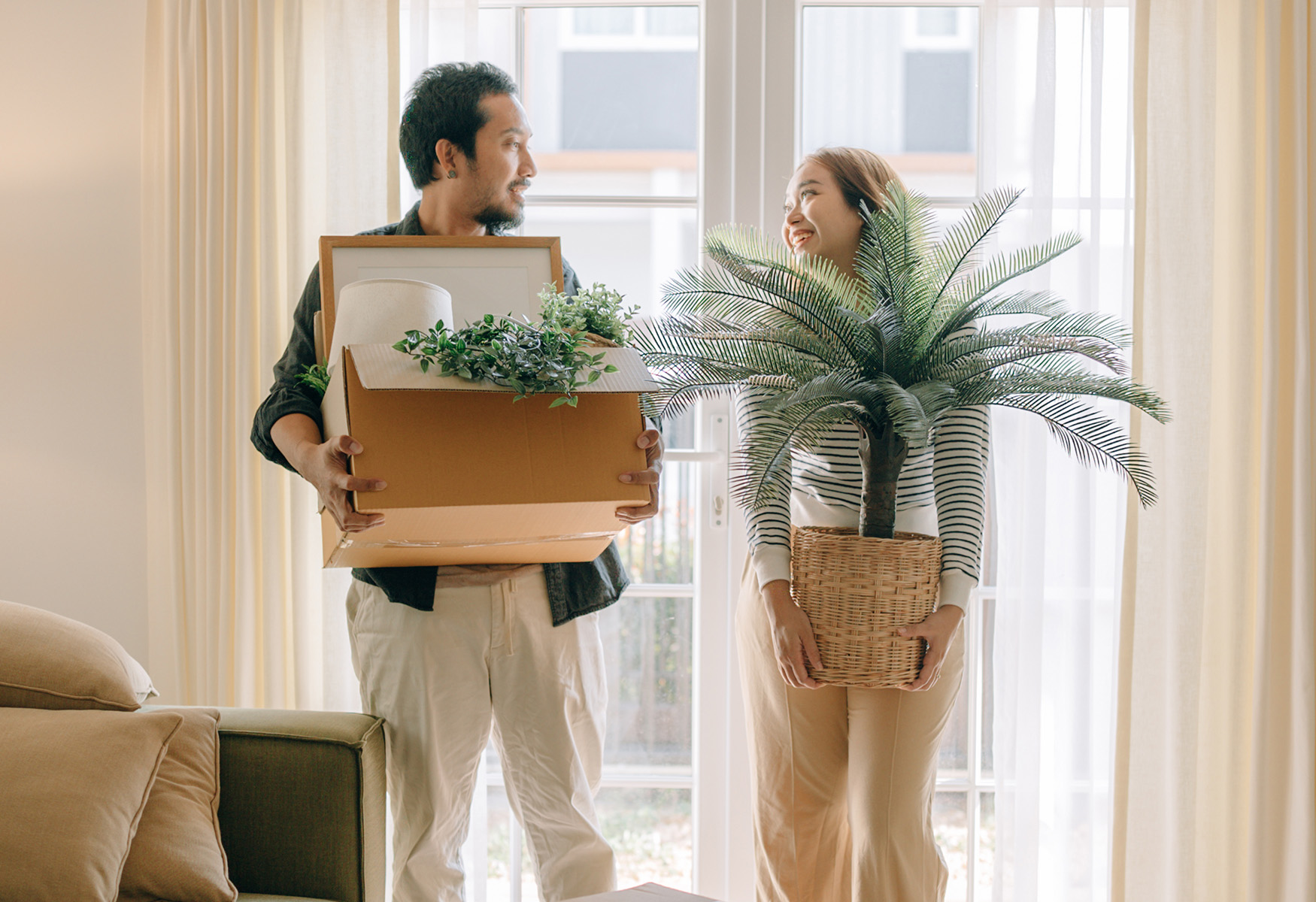 07-Section-7_IMG_01 A couple moving into a new home, one carrying a box of plants and décor, the other a potted palm